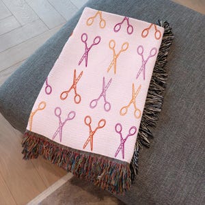 Op de afbeelding: Een lichtroze sprei met een herhalend patroon van scharen in oranje, paars en geel. De sprei heeft een gefranjerde rand met een mix van kleurrijke draden. Het is gedrapeerd over een grijze ondergrond.