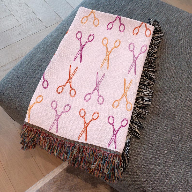Op de afbeelding: Een lichtroze sprei met een herhalend patroon van scharen in oranje, paars en geel. De sprei heeft een gefranjerde rand met een mix van kleurrijke draden. Het is gedrapeerd over een grijze ondergrond.