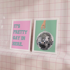 May include: Two framed prints on a white tiled wall. The print on the left has a pink background with the text "It's Pretty Gay In Here." in teal blue. The print on the right has a teal blue background with a silver disco ball hanging from a hand.