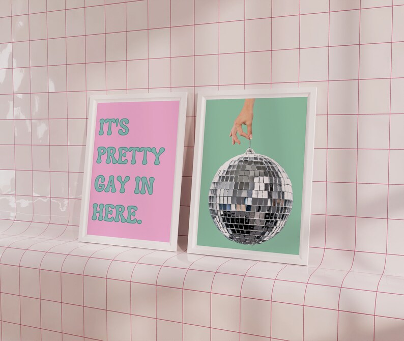 May include: Two framed prints on a white tiled wall. The print on the left has a pink background with the text "It's Pretty Gay In Here." in teal blue. The print on the right has a teal blue background with a silver disco ball hanging from a hand.