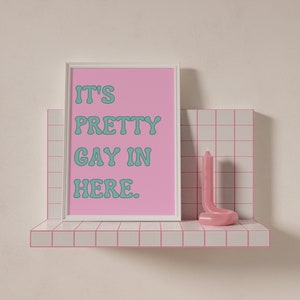 May include: A white framed print with pink background and teal text that reads "It's Pretty Gay In Here." The print is on a white shelf with pink tile and a pink candle.
