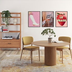 K&ouml;nnte beinhalten: Ein runder Esstisch aus Holz mit einem geriffelten Sockel. Zwei gelbe St&uuml;hle stehen auf beiden Seiten des Tisches. Der Tisch ist mit einem Buch und einer kleinen Topfpflanze gedeckt. An der Wand hinter dem Tisch h&auml;ngen drei gerahmte Drucke. Die Drucke zeigen einen rosa Hintergrund mit einem wei&szlig;en Stiefel, einer Discokugel und einer roten Erdbeere mit dem Text "LA FRAISE".
