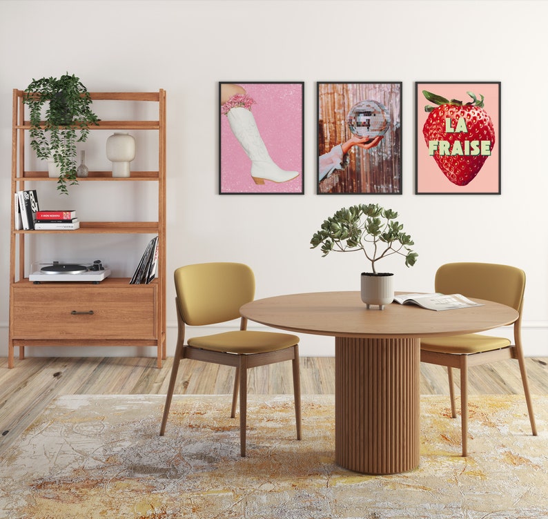 K&ouml;nnte beinhalten: Ein runder Esstisch aus Holz mit einem geriffelten Sockel. Zwei gelbe St&uuml;hle stehen auf beiden Seiten des Tisches. Der Tisch ist mit einem Buch und einer kleinen Topfpflanze gedeckt. An der Wand hinter dem Tisch h&auml;ngen drei gerahmte Drucke. Die Drucke zeigen einen rosa Hintergrund mit einem wei&szlig;en Stiefel, einer Discokugel und einer roten Erdbeere mit dem Text "LA FRAISE".