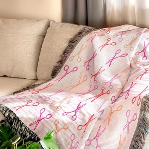 Op de afbeelding: Een witte sprei met een herhaald patroon van scharen in rood, oranje en roze tinten. De sprei heeft een gefranjerde rand en is over een beige bank gedrapeerd. Het scharenontwerp voegt een vleugje vrolijkheid toe.