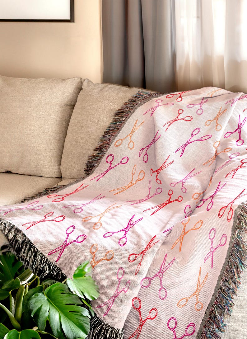 Op de afbeelding: Een witte sprei met een herhaald patroon van scharen in rood, oranje en roze tinten. De sprei heeft een gefranjerde rand en is over een beige bank gedrapeerd. Het scharenontwerp voegt een vleugje vrolijkheid toe.