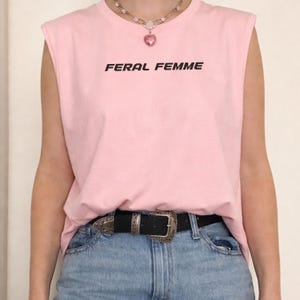 May include: Pink sleeveless shirt with the text "FERAL FEMME" in black. The shirt is paired with blue jeans and a black belt with a silver buckle. A necklace with a pink heart pendant is also visible.