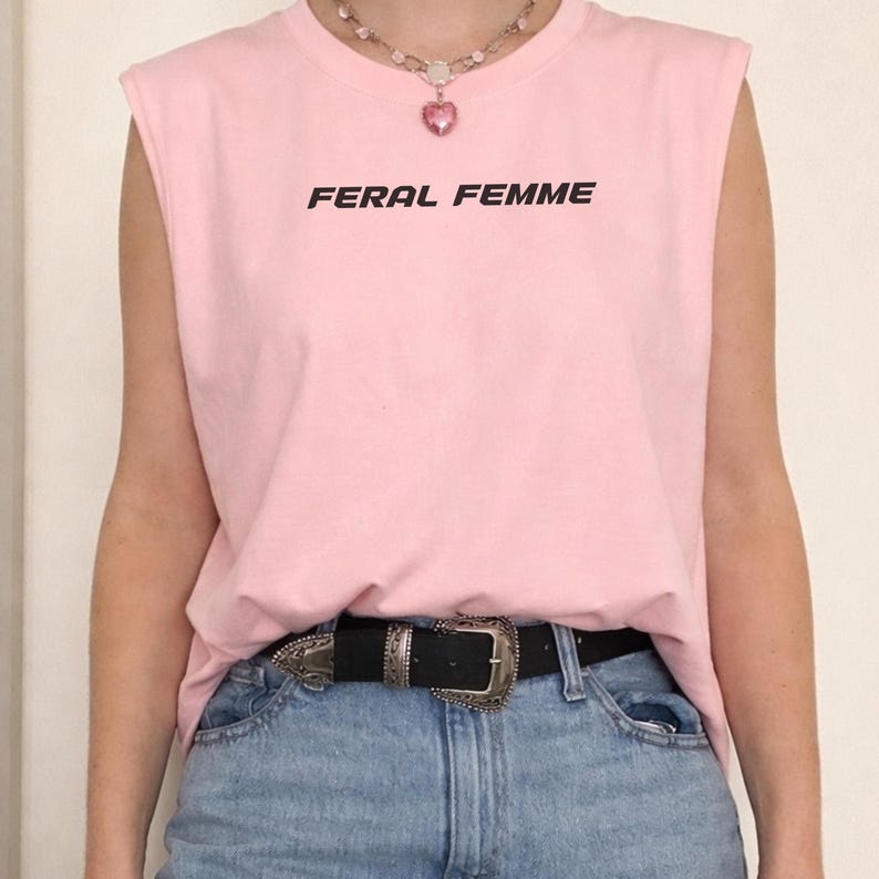 May include: Pink sleeveless shirt with the text "FERAL FEMME" in black. The shirt is paired with blue jeans and a black belt with a silver buckle. A necklace with a pink heart pendant is also visible.