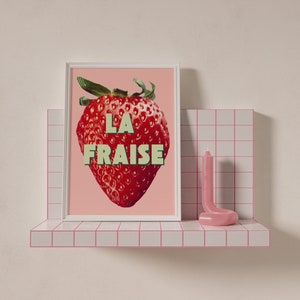 K&ouml;nnte beinhalten: Ein rosa gerahmter Druck mit einer roten Erdbeere und dem Text "LA FRAISE" in gr&uuml;nen und wei&szlig;en Buchstaben. Der Druck befindet sich auf einem wei&szlig;en Regal mit rosa Fliesen.
