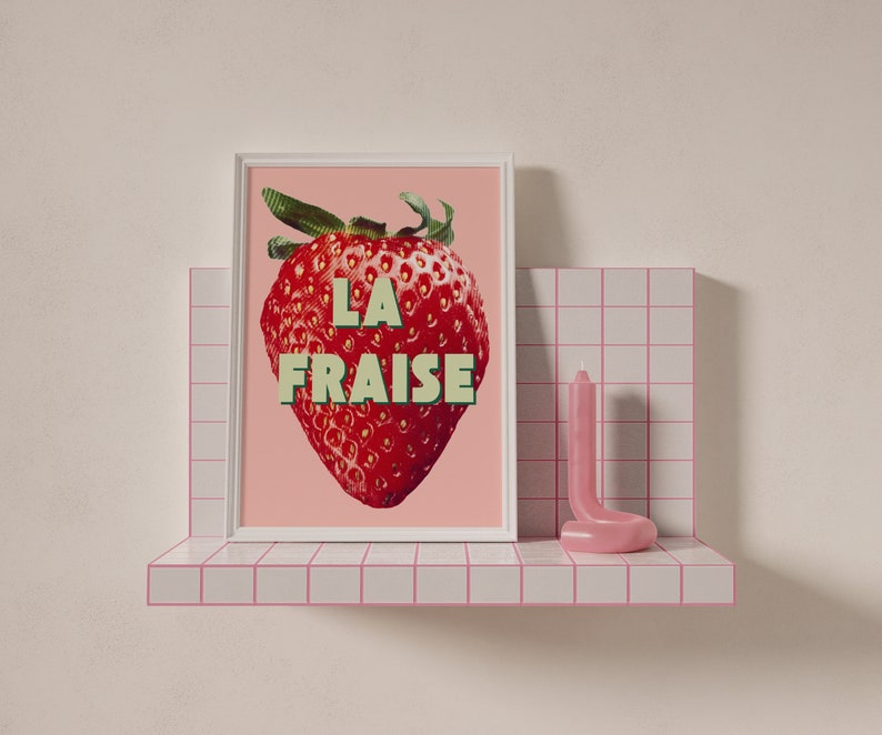 K&ouml;nnte beinhalten: Ein rosa gerahmter Druck mit einer roten Erdbeere und dem Text "LA FRAISE" in gr&uuml;nen und wei&szlig;en Buchstaben. Der Druck befindet sich auf einem wei&szlig;en Regal mit rosa Fliesen.