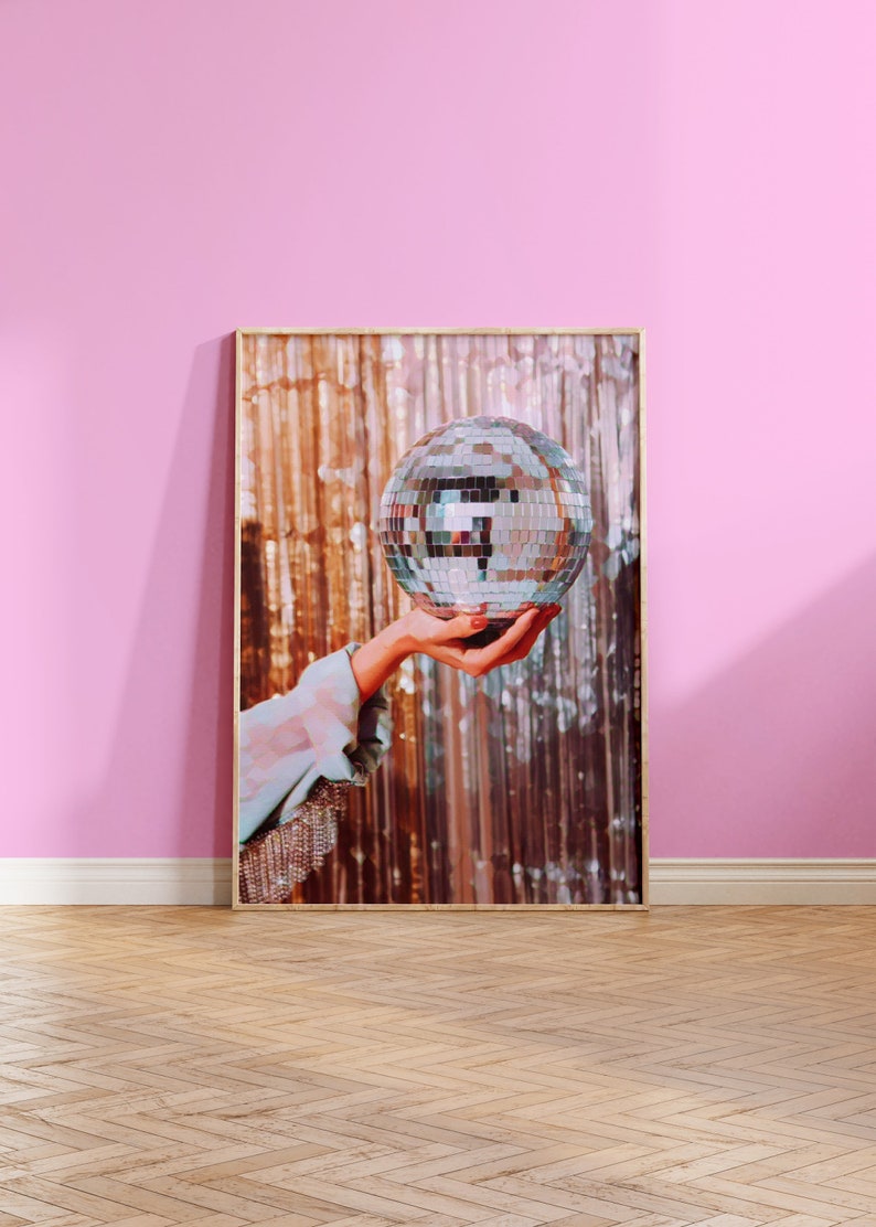 May include: A silver disco ball is held up in front of a shimmering gold background. The disco ball is reflecting light and has a mirrored surface.