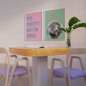 May include: Two framed posters hang on a white tiled wall above a wooden table with two purple chairs. The poster on the left says "It's Pretty Gay In Here." The poster on the right shows a hand holding a disco ball.