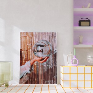 May include: A silver disco ball is held in a hand against a backdrop of shimmering gold and brown fabric. The disco ball is a classic party decoration.