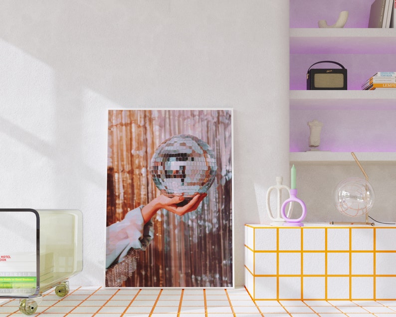 May include: A silver disco ball is held in a hand against a backdrop of shimmering gold and brown fabric. The disco ball is a classic party decoration.
