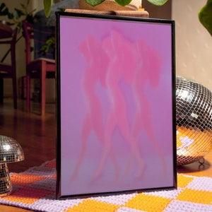 May include: A framed print with a pink and purple gradient background featuring three blurry silhouettes of people dancing. The frame is black and the print is sitting on a purple and orange checkered rug.
