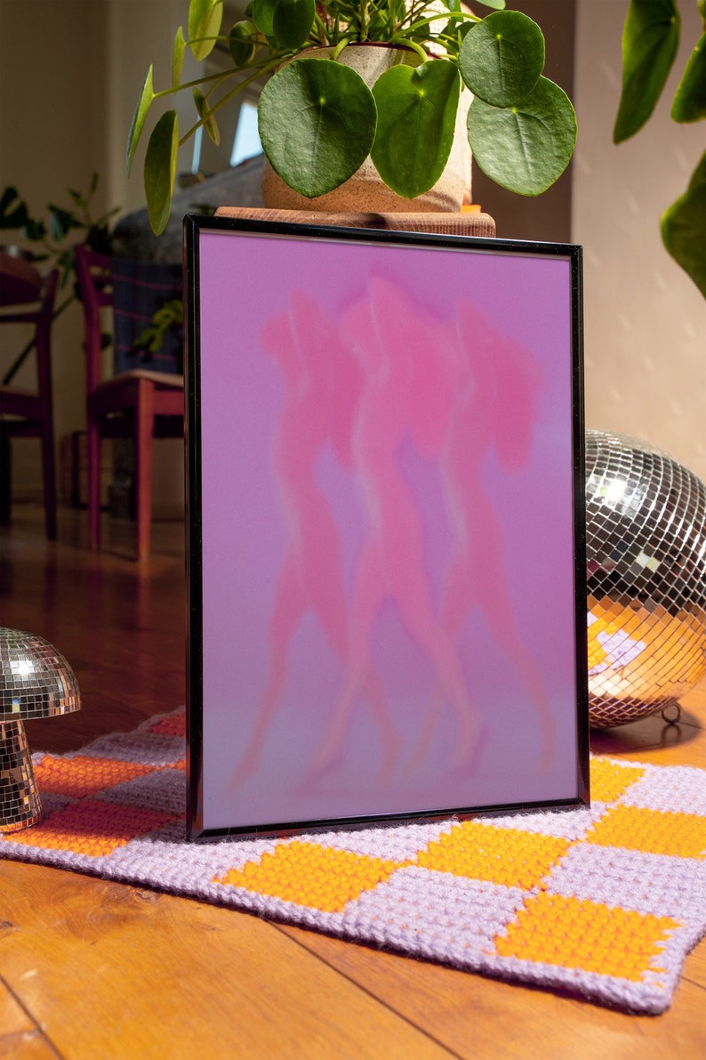 May include: A framed print with a pink and purple gradient background featuring three blurry silhouettes of people dancing. The frame is black and the print is sitting on a purple and orange checkered rug.