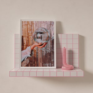 May include: A white picture frame with a photo of a hand holding a silver disco ball. The photo is on a pink tiled shelf with a pink candle.