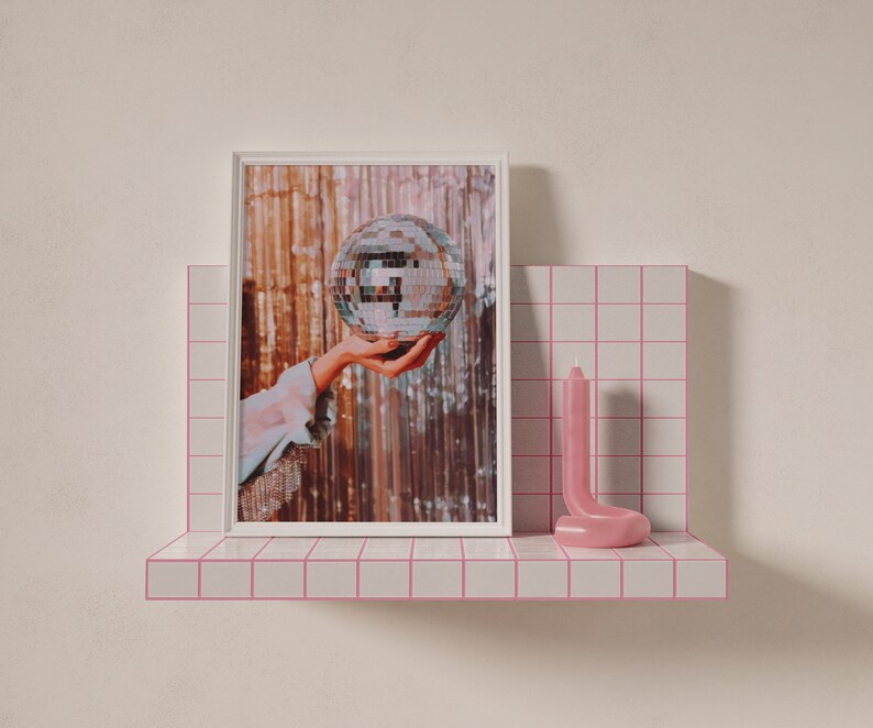 May include: A white picture frame with a photo of a hand holding a silver disco ball. The photo is on a pink tiled shelf with a pink candle.