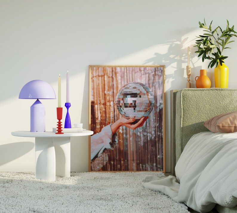 May include: A white bedside table with a purple lamp and three candlesticks. The candlesticks are red, blue, and white. The table is in front of a bed with a white duvet and a green headboard. A framed print of a hand holding a disco ball is on the wall behind the table.