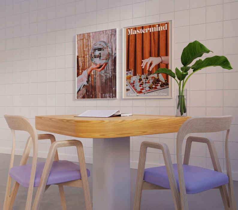 May include: Two wooden chairs with purple cushions and a wooden table with a book on top. The table has a white base. Two framed posters hang on a white tiled wall behind the table. The posters feature a disco ball and a chessboard with the text "Mastermind. Checkmate. I couldn't lose."