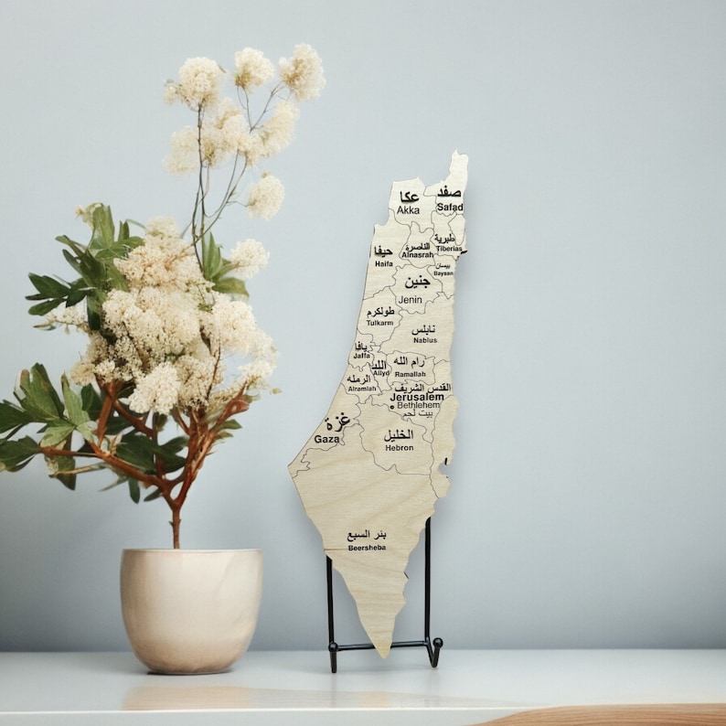 May include: A wooden map of Israel with the names of cities and towns in Arabic script. The map is on a black metal stand and is in front of a white wall.
