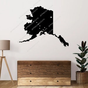 Può includere: Una silhouette nera dello stato dell'Alaska con stelle bianche su una parete bianca. La silhouette è su una parete con una cassettiera in legno e una pianta in vaso in primo piano.