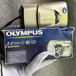 May include: A silver Olympus mju: III 120 camera with a black wrist strap, resting on its original blue and white box. The box features the text Olympus mju: III 120 Quartzdate, 38 mm-120 mm Zoom, Multi-Wide AF System, Camera Shake Indicator, and All-weather construction.