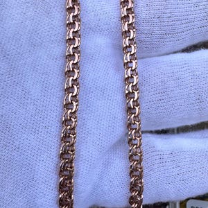 May include: Close-up of two rose gold chain necklaces with a textured, interlocking design. The chains are displayed on a white surface, with a scale visible in the background. The chains are a warm, metallic color.