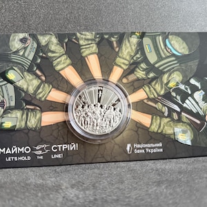 May include: A silver coin with a design of soldiers standing shoulder to shoulder. The coin is surrounded by a cartoon image of soldiers holding hands, with the text "Тримаймо стрій!" and "Let's hold the line!" at the bottom. The text "Національний банк України" is also visible.