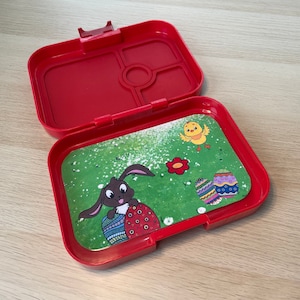 Könnte beinhalten: Rote Plastik-Lunchbox mit einem herausnehmbaren Tablett mit einem Cartoon-Hasen, der ein Osterei hält, einem Küken und einem Osterei-Design. Das Tablett ist grün mit weißen Flecken.