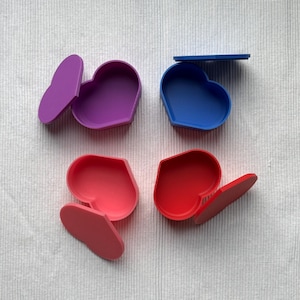 Könnte beinhalten: Vier herzförmige Boxen mit Deckeln, jede in einer anderen Farbe: lila, blau, rosa und rot.