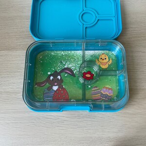 K&ouml;nnte beinhalten: Eine blaue Plastik-Lunchbox mit einem durchsichtigen Deckel. Der Deckel hat ein gr&uuml;nes, grasartiges Design mit einem Hasen, der Ostereier h&auml;lt, und einem K&uuml;ken. Die Lunchbox hat vier F&auml;cher.