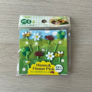 May include: A pack of 8 colourful plastic food picks with bee, ladybug, clover, and flower designs. The pack says "Honey & Flower Pick" and "8本入 4種類×2本".