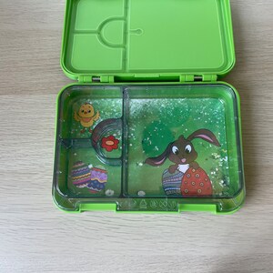 K&ouml;nnte beinhalten: Gr&uuml;ne Lunchbox mit durchsichtigem Deckel. Die Lunchbox hat vier F&auml;cher. Die F&auml;cher haben Bilder von einem K&uuml;ken, einer Blume, einem Hasen, der ein Ei h&auml;lt, und einem gr&uuml;n-wei&szlig;en Ei.