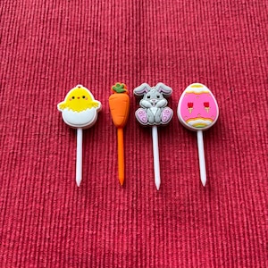 May include: Four Easter-themed cake toppers: a yellow chick, a carrot, a gray bunny, and a pink and white egg. Each topper has a white stick.