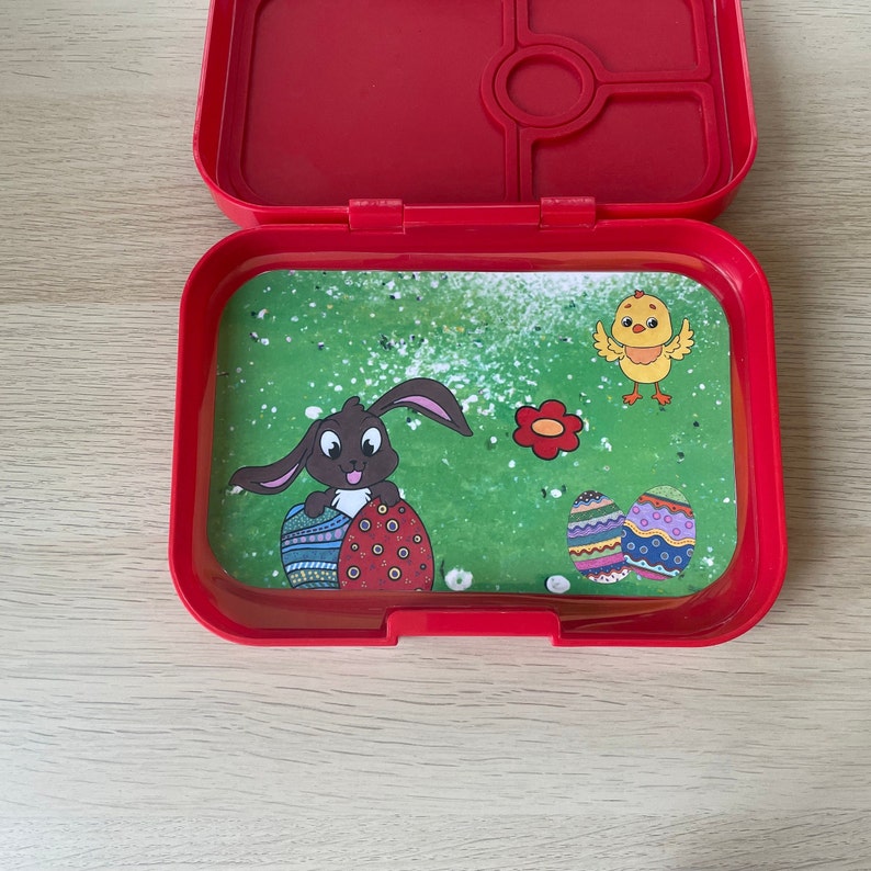 K&ouml;nnte beinhalten: Rote Lunchbox mit einem gr&uuml;n-wei&szlig;-braunen Ostermotiv. Das Motiv zeigt ein H&auml;schen, das Ostereier h&auml;lt, ein K&uuml;ken und Blumen.