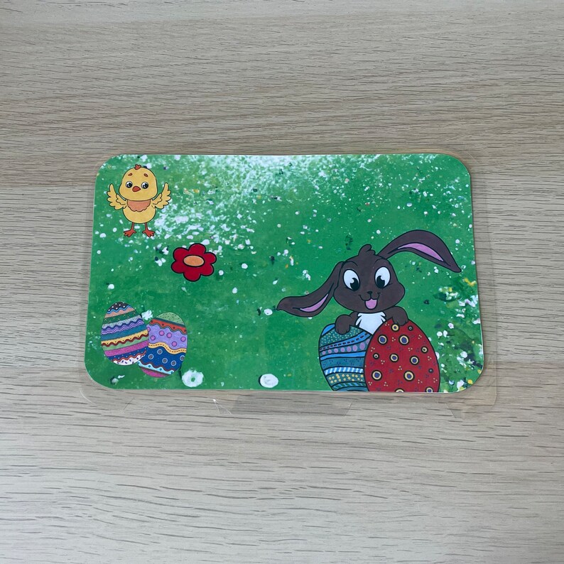 K&ouml;nnte beinhalten: Ein gr&uuml;nes und wei&szlig;es Oster-Platzset mit einem braunen Hasen, der zwei verzierte Eier h&auml;lt. Ein gelbes K&uuml;ken mit roten F&uuml;&szlig;en befindet sich oben links.