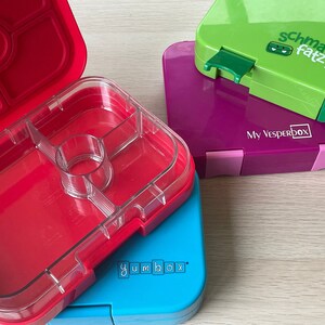 K&ouml;nnte beinhalten: Vier bunte Lunchboxen, die &uuml;bereinander gestapelt sind. Die oberste Box ist gr&uuml;n mit dem Text "schmatz fatz junior" und einem gr&uuml;nen Monster-Logo. Die zweite Box ist lila mit dem Text "My Vesperbox". Die dritte Box ist blau mit dem Text "yumbox". Die unterste Box ist rot mit einem durchsichtigen Deckel und mehreren F&auml;chern.