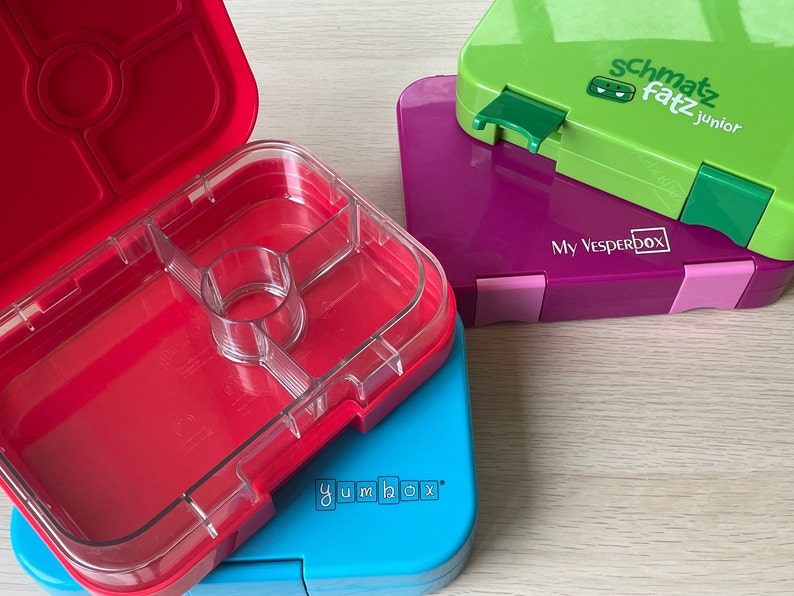 K&ouml;nnte beinhalten: Vier bunte Lunchboxen, die &uuml;bereinander gestapelt sind. Die oberste Box ist gr&uuml;n mit dem Text "schmatz fatz junior" und einem gr&uuml;nen Monster-Logo. Die zweite Box ist lila mit dem Text "My Vesperbox". Die dritte Box ist blau mit dem Text "yumbox". Die unterste Box ist rot mit einem durchsichtigen Deckel und mehreren F&auml;chern.
