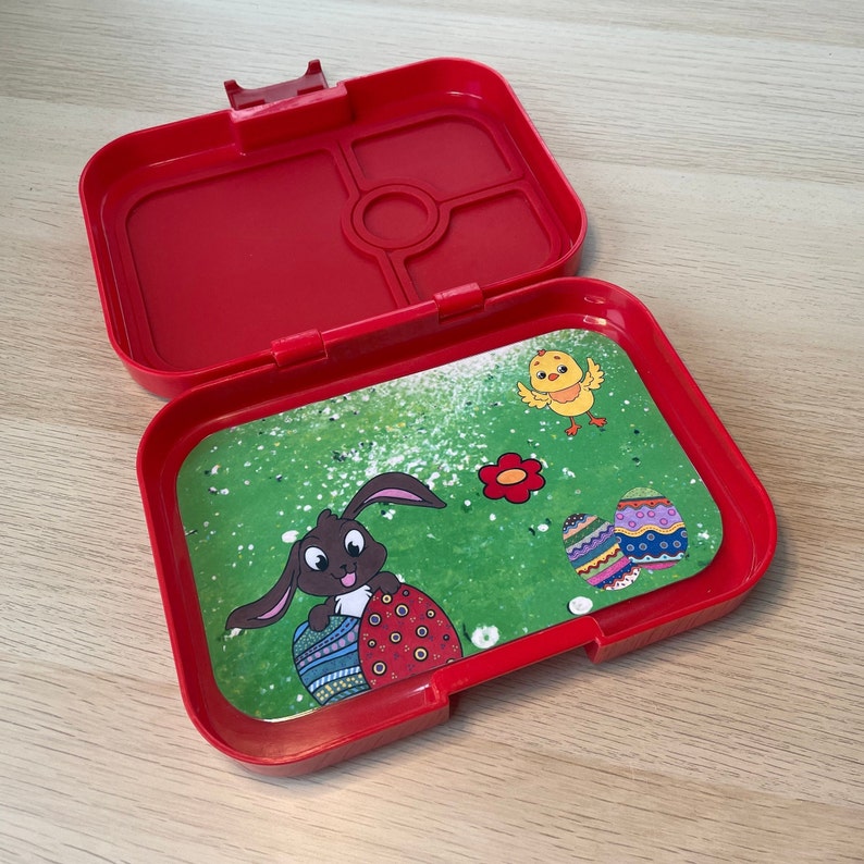 K&ouml;nnte beinhalten: Rote Plastik-Lunchbox mit einem herausnehmbaren Tablett mit einem Cartoon-Hasen, der ein Osterei h&auml;lt, einem K&uuml;ken und einem Osterei-Design. Das Tablett ist gr&uuml;n mit wei&szlig;en Flecken.