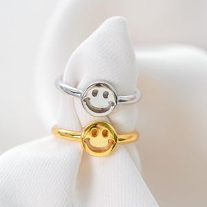 May include: Two smiley face rings, one silver and one gold, on a white fabric background.