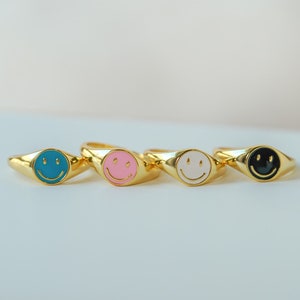 May include: Four gold signet rings with different colored enamel smiley faces. The rings are teal, pink, white, and black.