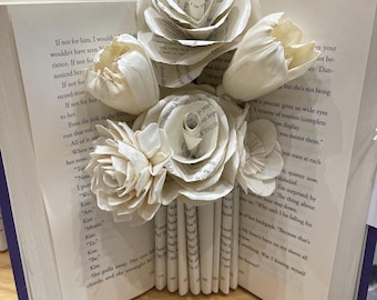 May include: A decorative book with pages folded to resemble a bouquet of white paper flowers. The book is open, revealing printed text, and rests on a wooden surface. The flowers are clustered at the top of the book.