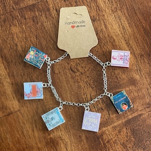 May include: A silver charm bracelet with six charms featuring miniature book covers. The charms include Pride and Prejudice, Emma, Persuasion, Sense and Sensibility, Little Women, and a book with a floral design.