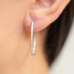 Peut inclure: Gros plan d'une boucle d'oreille créole argentée ornée d'une rangée de petites pierres scintillantes. La boucle d'oreille est portée sur une oreille, avec la peau et les cheveux de la personne visibles en arrière-plan. Bijou élégant.