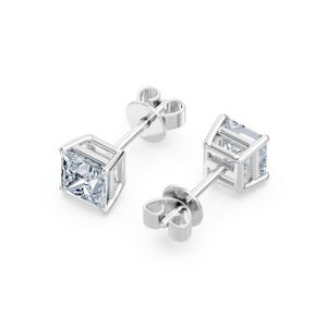 May include: A pair of silver-toned stud earrings featuring square-cut clear stones set in a four-prong basket setting. Each earring includes a post back and a matching metal butterfly clutch closure, showcasing a clean and reflective metallic finish.