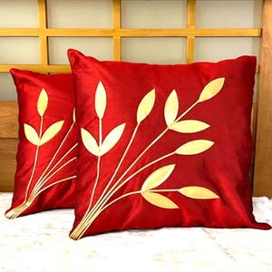 May include: Two red decorative throw pillows with a gold leaf design. The pillows are square-shaped and made of a shiny fabric. The gold leaf design is embroidered on the front of the pillows. The pillows are placed on a white surface.