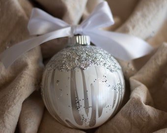 May include: A white Christmas ornament with silver glitter accents and vertical silver drip designs. The ornament is topped with a white satin bow and ribbon. The background is a neutral-toned fabric.
