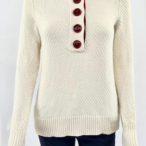 May include: Cream-colored sweater with a textured pattern, featuring a burgundy collar and button placket. The long-sleeved top has ribbed cuffs and hem, with four burgundy buttons down the front.