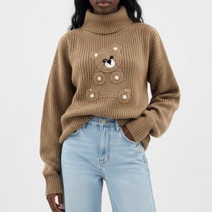 May include: A tan turtleneck sweater with a teddy bear design on the front. The teddy bear is made of a lighter brown material with white accents. The sweater has a ribbed knit texture and long sleeves.