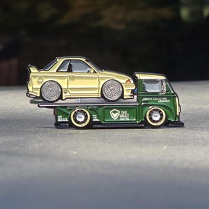 May include: A metal enamel pin featuring a cream-colored sports car loaded onto a dark green flatbed tow truck. The truck side displays the text "PRIVATE COLLECTION LOGISTICS" and "THE VAULT" with a shield logo. The wheels have detailed silver rims.